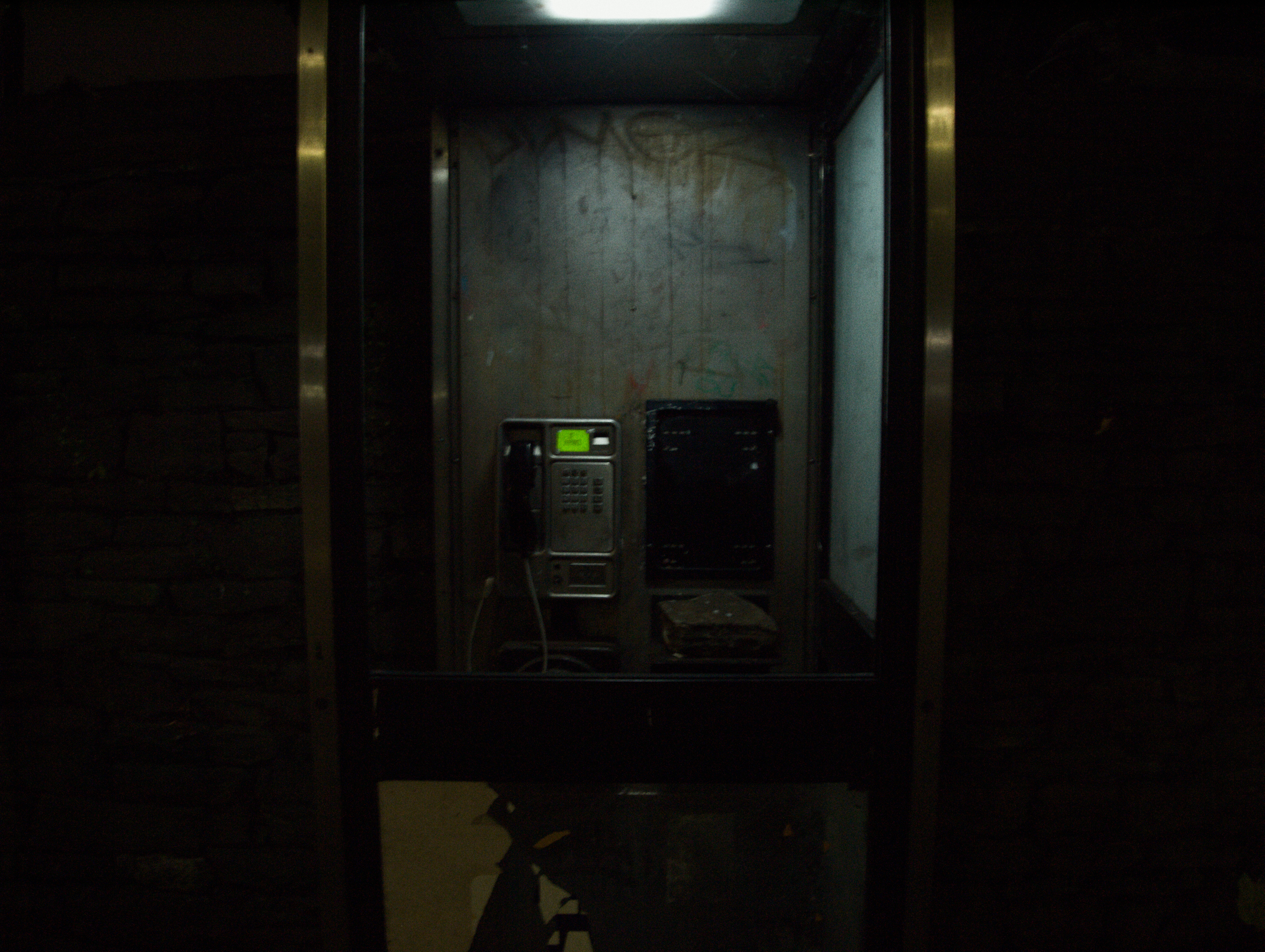 A dark grimy picture of a phonebox at night, a large block of stone of unknown origin sits on the little shelf where a phonebook once would have sat